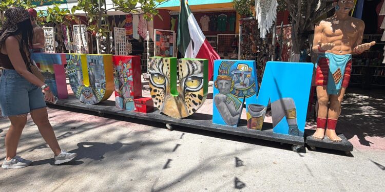 Letras gigantes de TULUM decoradas con motivos mayas junto a una estatua de un guerrero y la bandera de México